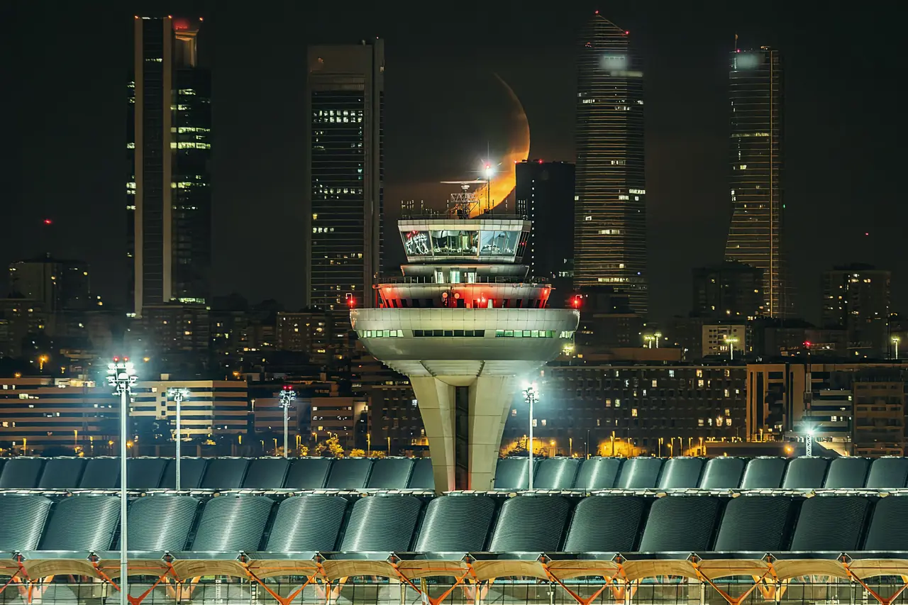 La torre de control y la cubierta ondulada de la T4 de Barajas de noche, con las Cuatro Torres y la luna creciente detrás
