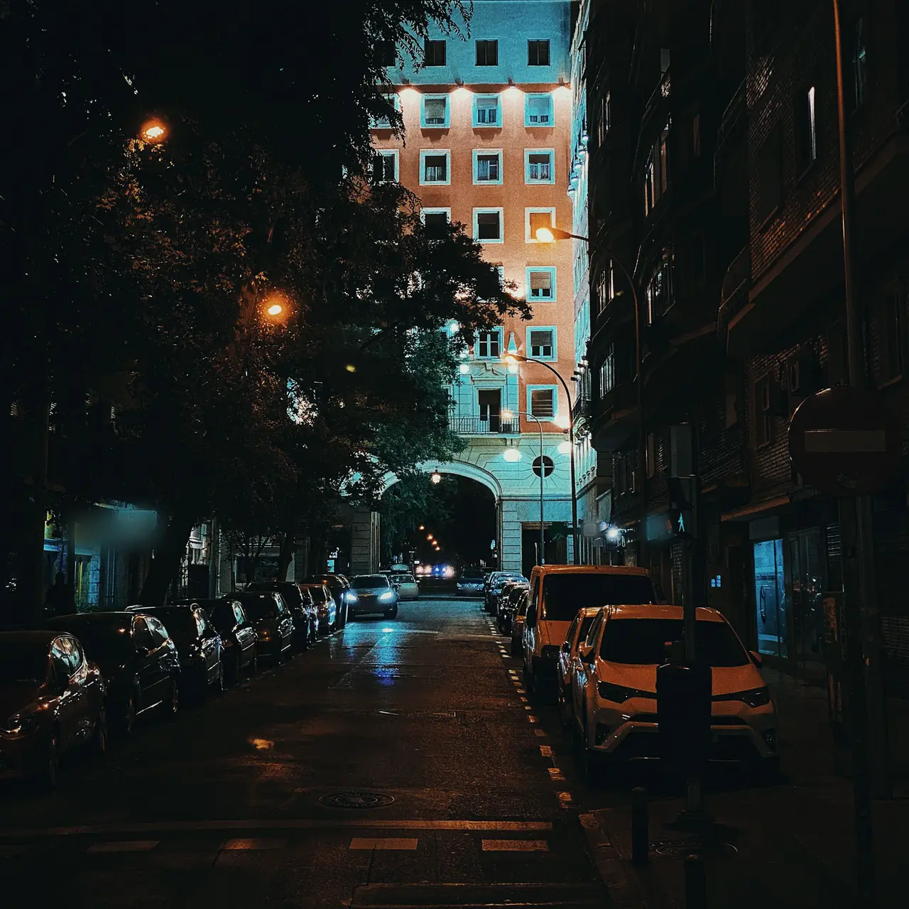 Calle de Madrid de noche con coches aparcados a los dos lados, un SUV blanco junto al bordillo y farolas de sodio